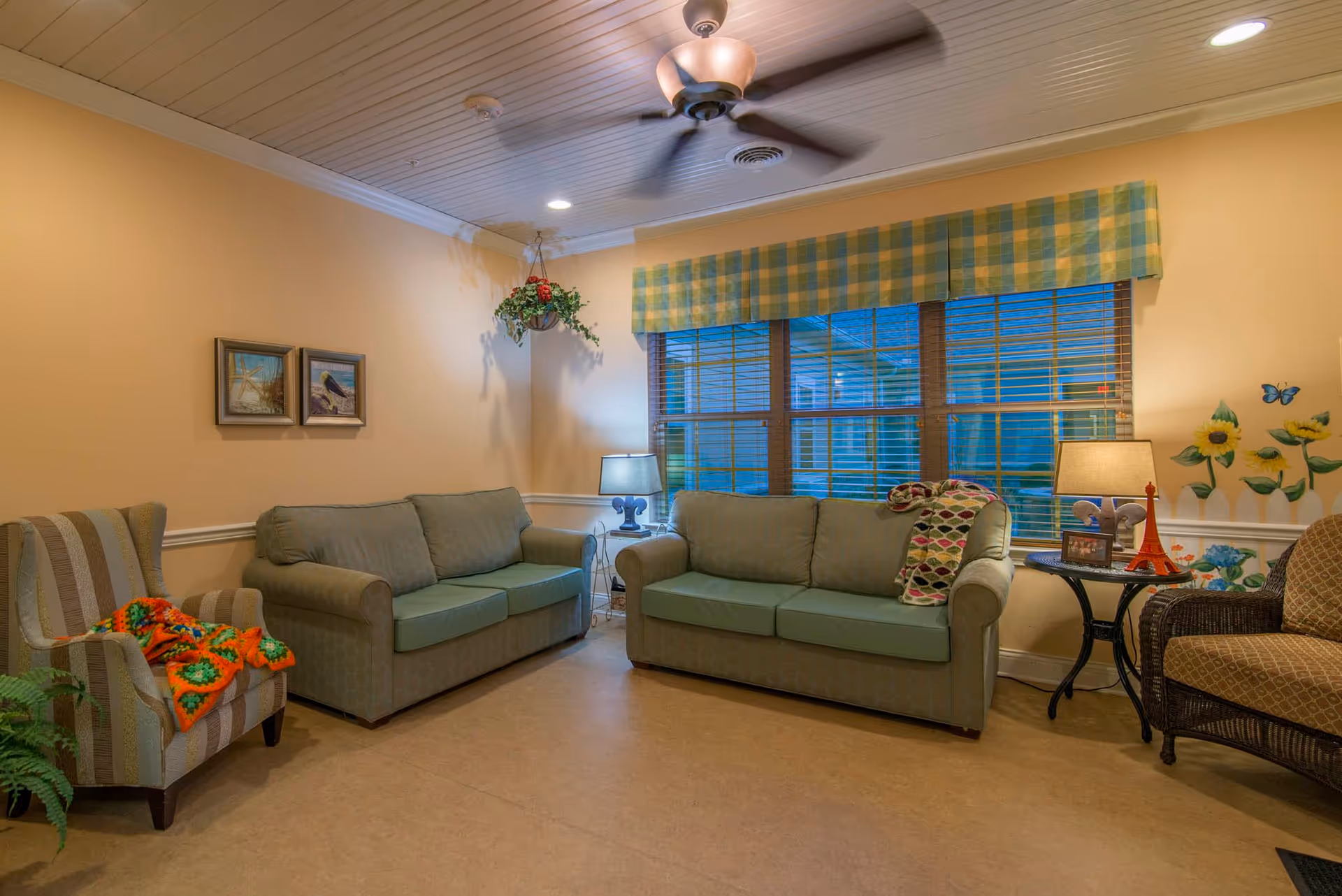 A cozy living room with two green sofas, a striped armchair with a colorful blanket, a wicker chair, and a small round table with a lamp and decorative items. The room has beige walls, a ceiling fan, a large window with yellow and green plaid valance, and wall decorations including framed pictures and sunflower decals.