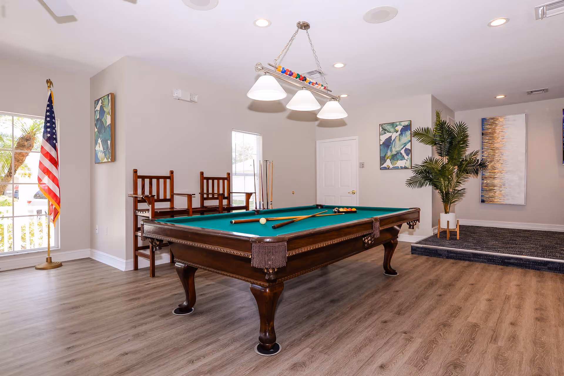 A well-lit indoor recreational room featuring a classic wooden pool table with green felt, two pool cues resting on the table, and a rack of billiard balls hanging from a ceiling light fixture. The room has light-colored walls adorned with abstract artwork, a large window with an American flag beside it, two wooden chairs against the wall, a potted plant on a small raised platform, and wood-style flooring.