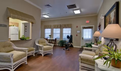 A cozy living room area with wicker armchairs and a loveseat with beige cushions arranged around a small table. The room has wood flooring, a few potted plants, a lamp on a side table, and a window with beige valances. There is an exercise bike near the back door, and a small pass-through window to a kitchen area with a refrigerator visible.