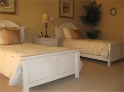 A bedroom with two white twin beds, each with beige bedding and a brown pillow. Between the beds is a white nightstand with a lamp and a small framed photo. The walls are beige and decorated with framed artwork. There is a green potted plant in the corner of the room.