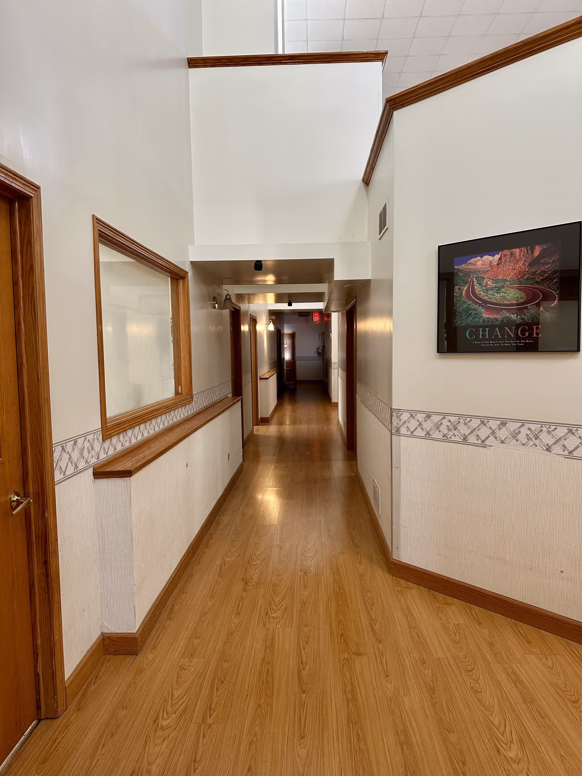 A long, narrow hallway with wooden flooring and white walls trimmed with wood. There are several doors along the hallway and a large window on the left wall. A framed picture with the word 'CHANGE' is hung on the right wall. The hallway is well-lit with ceiling lights and has a high ceiling at the near end.
