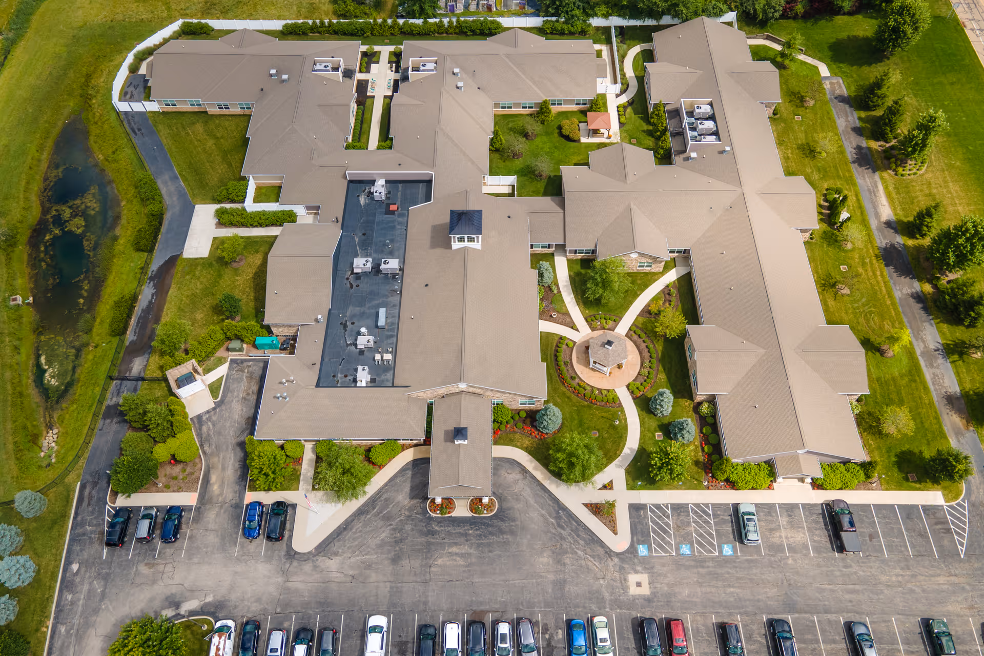 Aerial view of Renaissance of Richfield/Bath senior living facility showing a large building with multiple wings, surrounded by landscaped green areas, pathways, parking lots with cars, and a small pond on the left side.