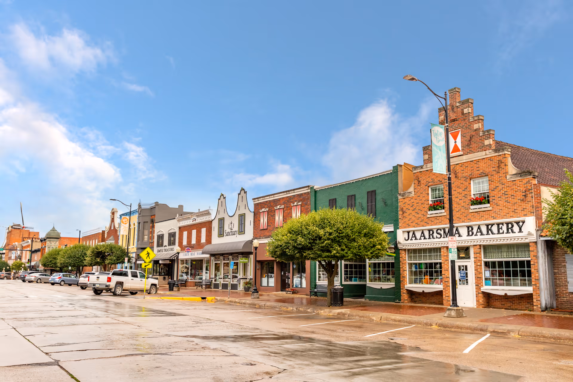 Street view of a small town with a row of colorful historic buildings including Jaarsma Bakery, trees lining the sidewalk, parked cars, and a partly cloudy blue sky.