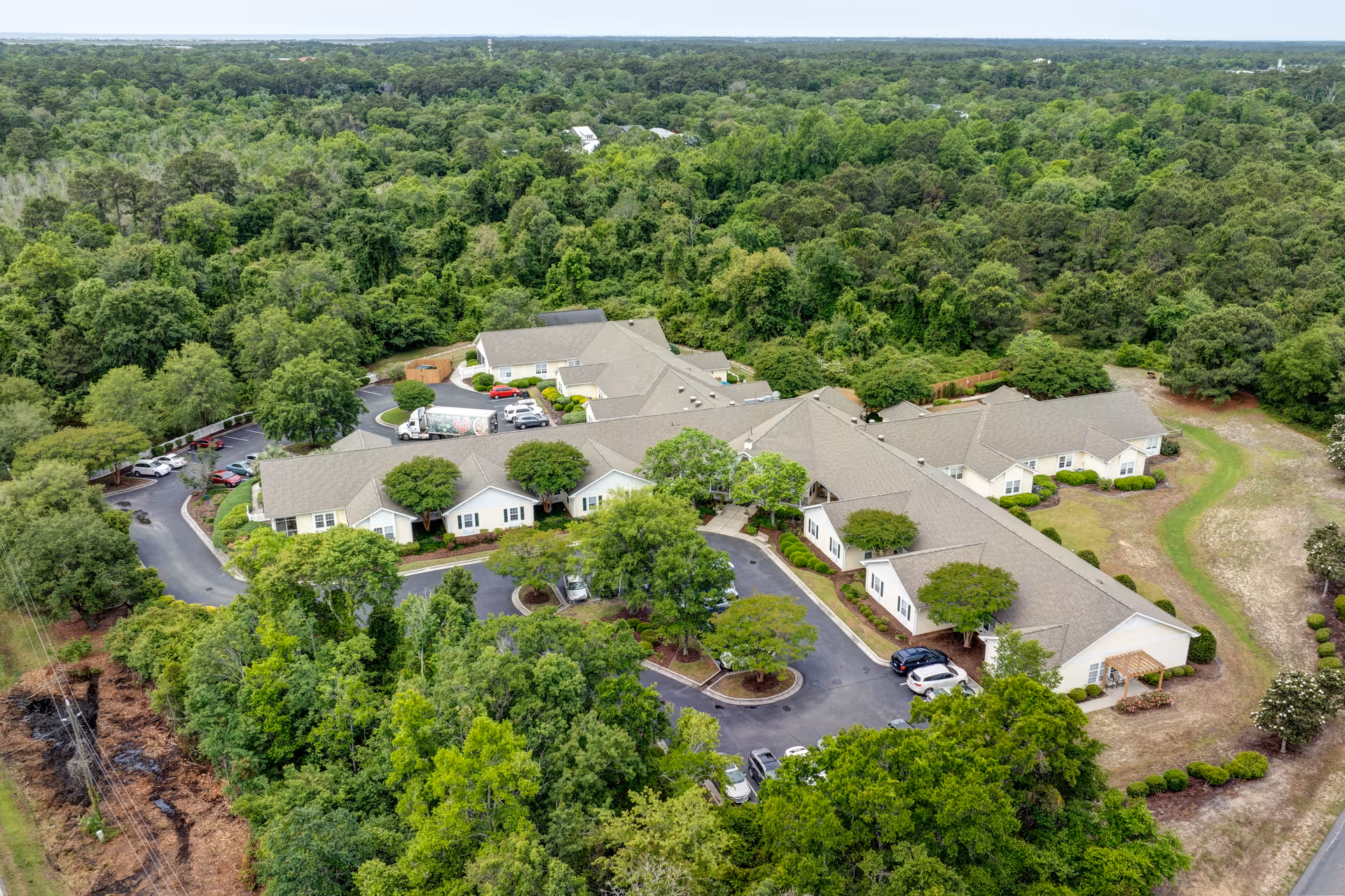 Aerial view of TerraBella Southport facility surrounded by dense green trees and vegetation. The building complex has multiple connected sections with beige roofs and white walls. There are several parking spaces with cars parked, and paved driveways winding through the property.
