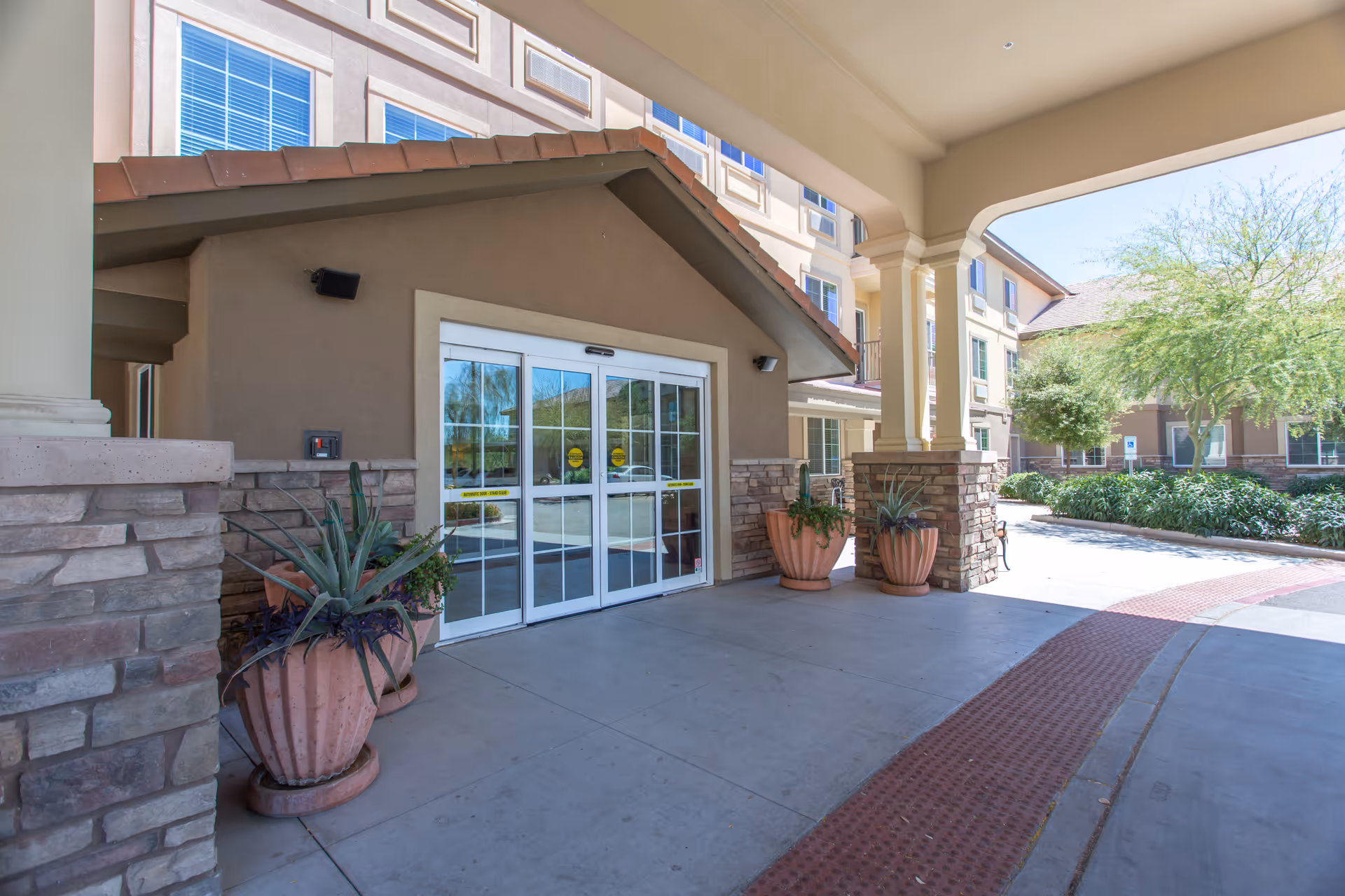 Entrance to a senior living facility with automatic sliding glass doors, large potted plants on either side, stone and stucco exterior walls, and a covered driveway area with columns. Trees and shrubs are visible in the background under a clear sky.