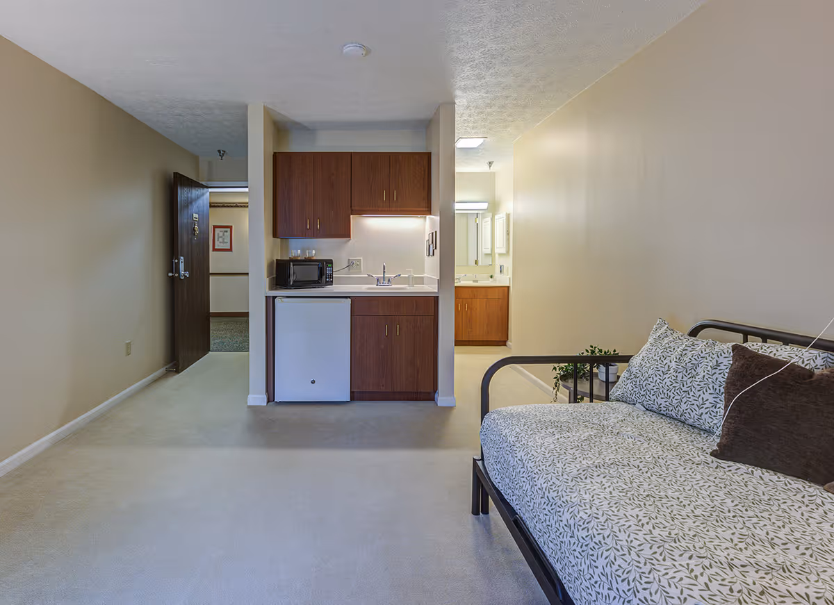Interior view of a senior living facility room showing a small kitchenette with a microwave, mini fridge, and sink. To the right, there is a daybed with patterned cushions. In the background, a bathroom vanity with a mirror is visible. The walls are painted beige and the floor is carpeted.