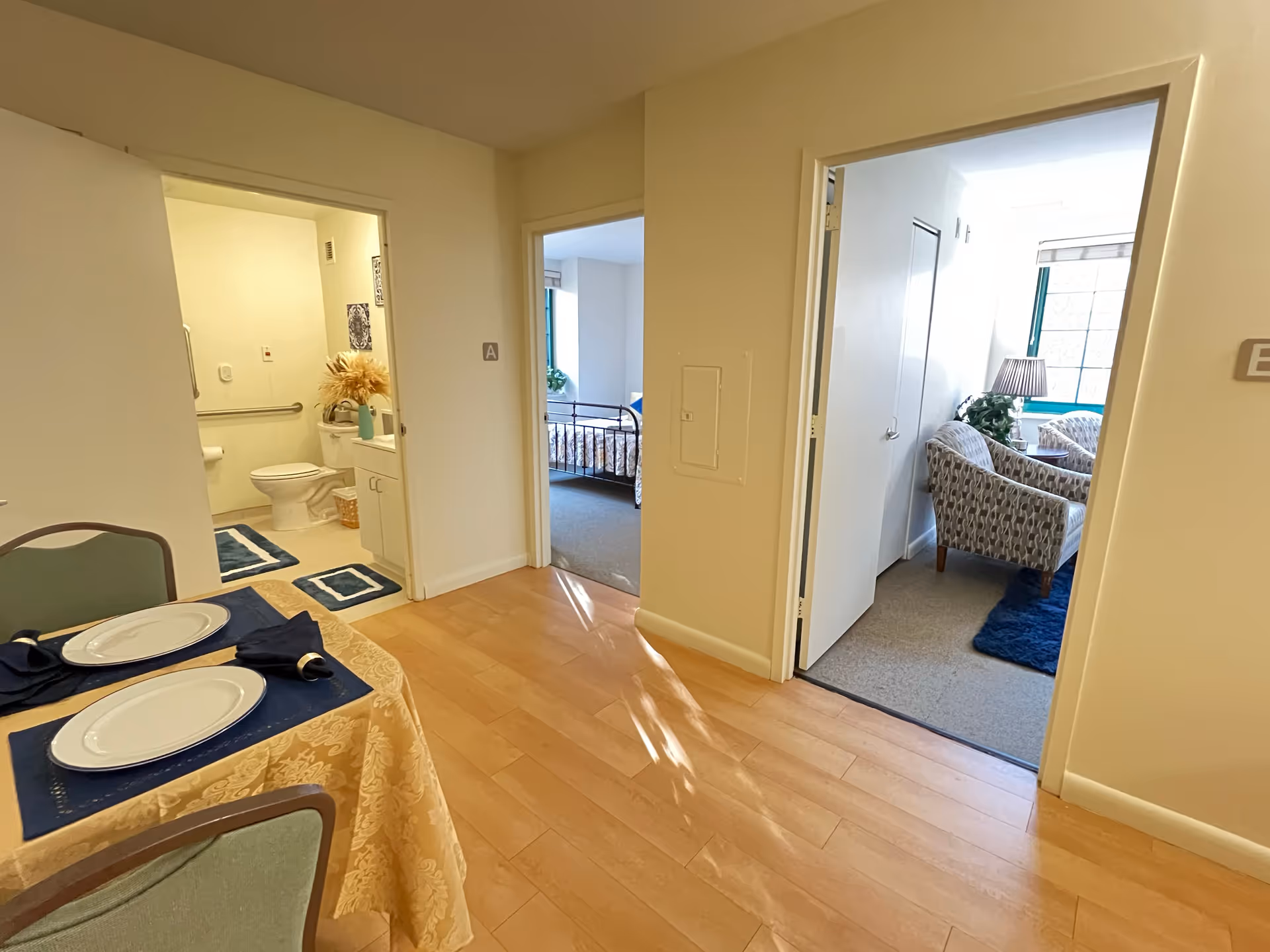 Interior view of a senior living apartment with a set dining table, an open bathroom, a bedroom doorway, and a sitting area.