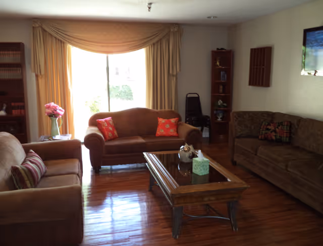 A cozy living room with three brown sofas arranged around a wooden coffee table with a glass top. The coffee table has a decorative item and a tissue box on it. There is a large window with beige curtains letting in natural light. The room has wooden flooring and shelves with books and decorative items.