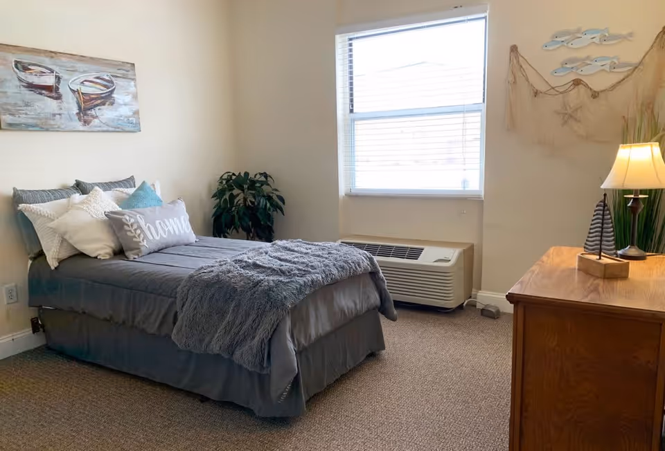 Furnished single bedroom with a made bed, decorative pillows, window air conditioner, and a dresser with a lamp.