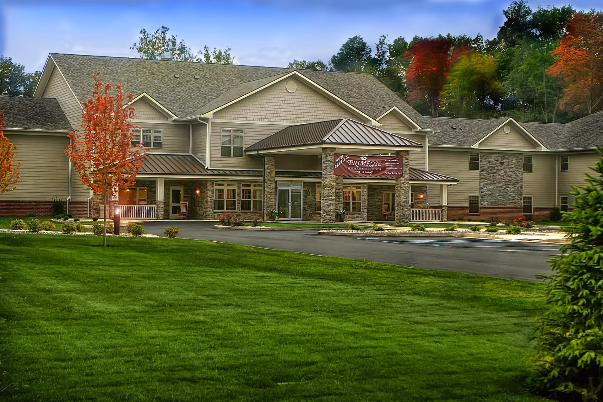 Exterior view of Primrose Retirement Community of Kokomo building with a large green lawn in the foreground, trees with autumn foliage, and a covered entrance with stone pillars and a sign displaying the facility name.