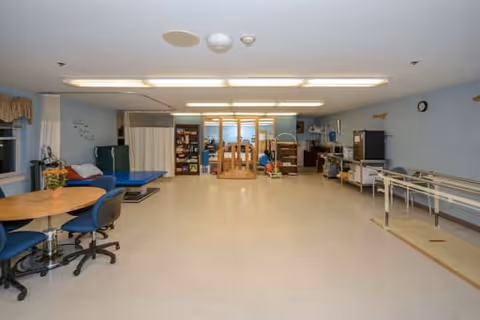 A spacious room with light blue walls and a beige floor, featuring several tables with blue chairs on the left, medical or therapy equipment including a treatment table and parallel bars on the right, and shelves with supplies and equipment at the back under bright fluorescent ceiling lights.