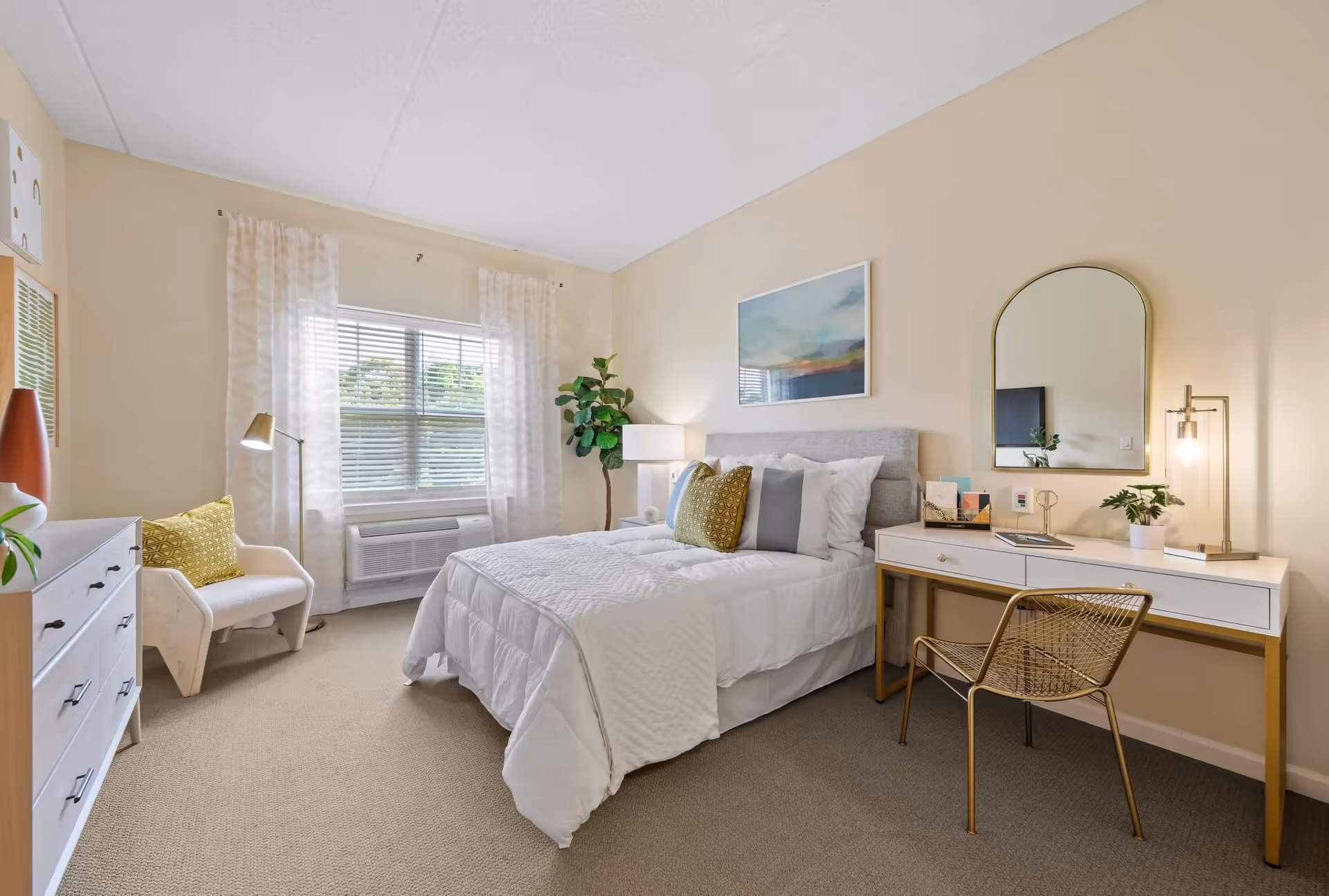 A bright and cozy bedroom with a neatly made bed featuring white bedding and multiple pillows. There is a white dresser with a plant and decorative items on the left, a white armchair with a yellow cushion near a window with sheer curtains, and a desk with a gold frame and a matching chair on the right. The desk has a lamp, a small plant, and some books. A large mirror hangs above the desk, and a framed artwork is on the wall above the bed. The room has beige walls and carpeted flooring.