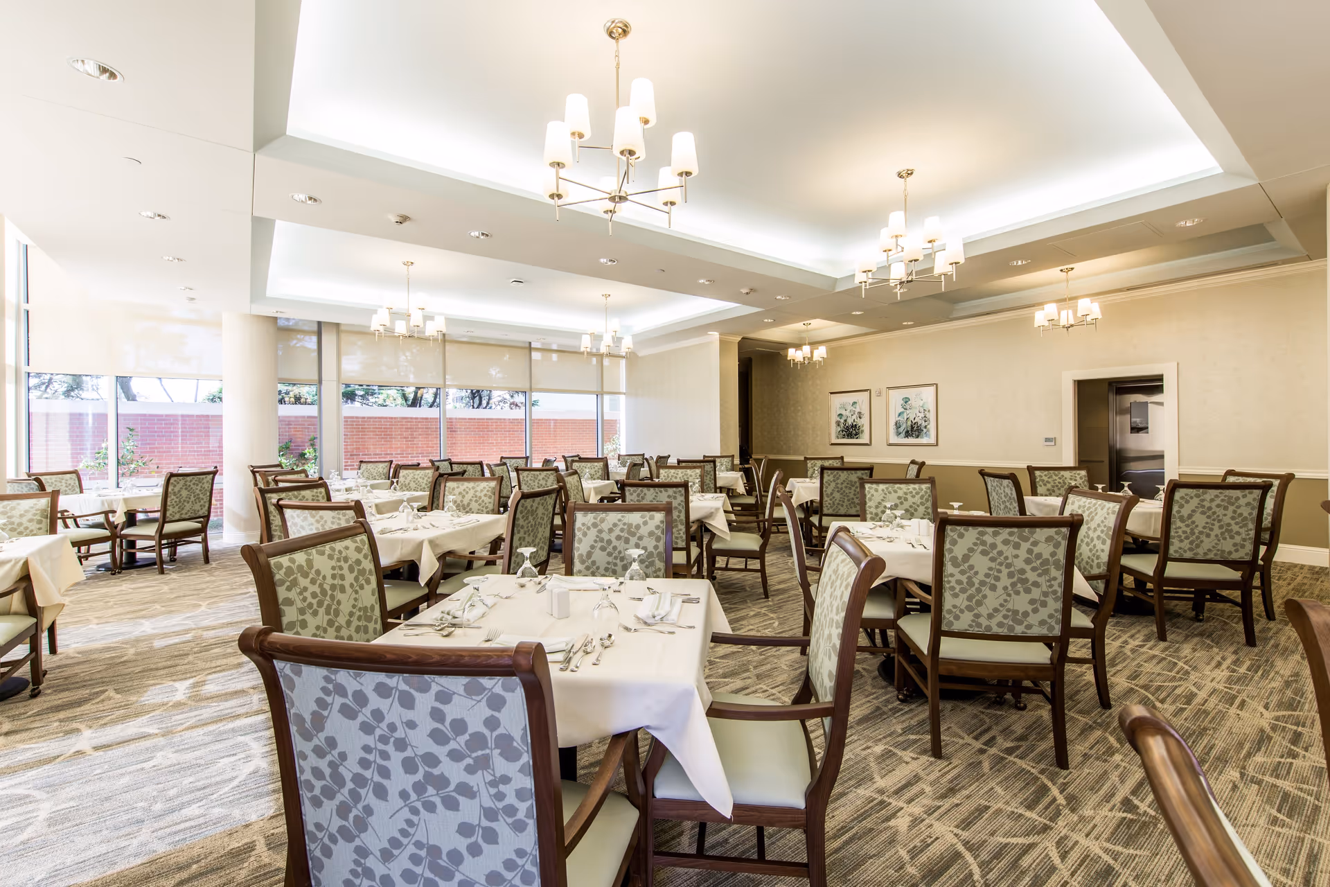 A bright and spacious dining room with multiple tables covered in white tablecloths, each set with silverware, napkins, and glasses. The chairs have wooden frames with patterned upholstery. Large windows on one side let in natural light, and elegant chandeliers hang from the ceiling. The room has a neutral color palette with beige walls and carpeted floors.
