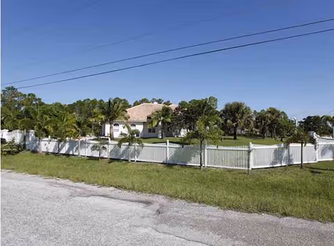 Exterior view of a fenced, grassy property with a house and trees under a clear sky.