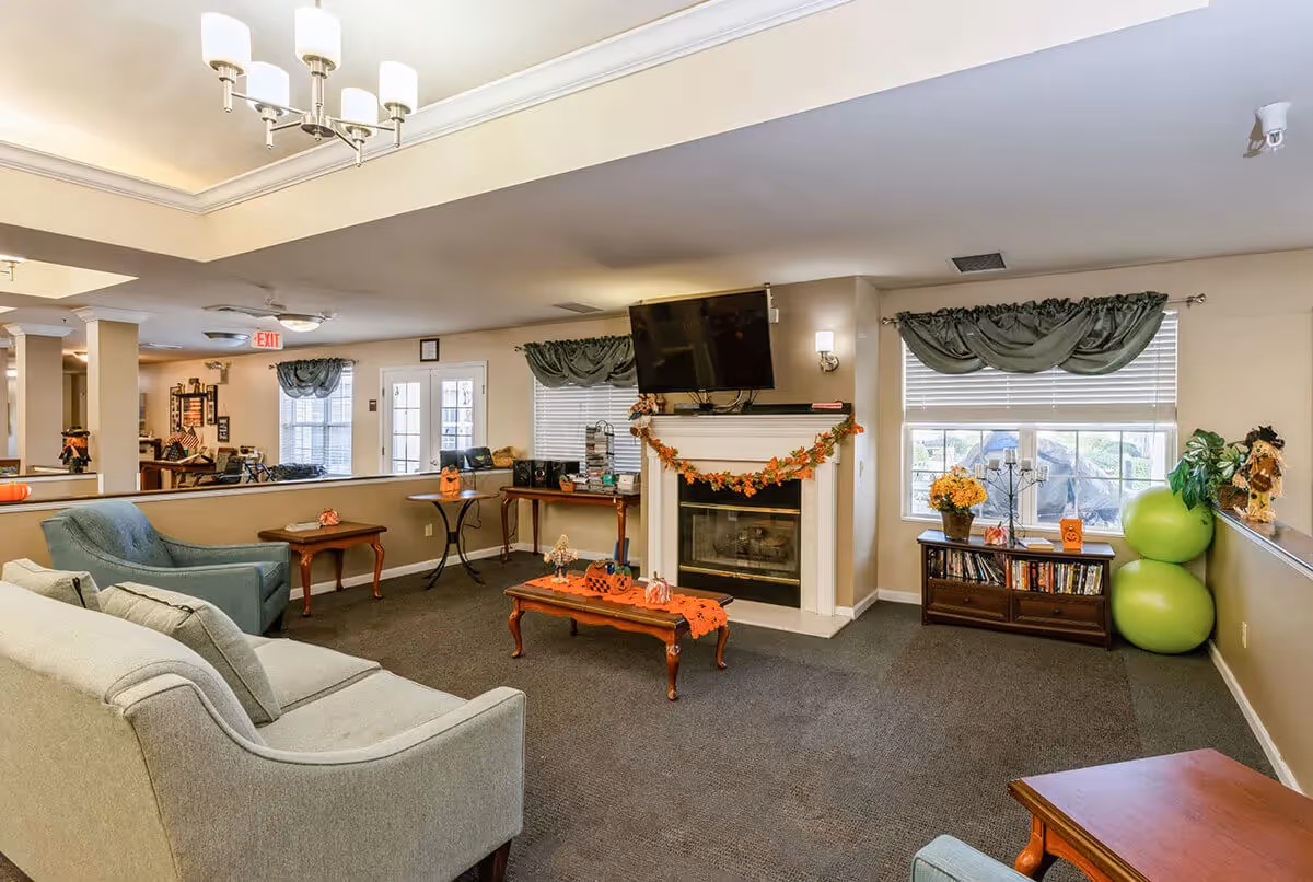 A cozy living room area in a senior living facility with a gray sofa and armchair, a wooden coffee table decorated with Halloween-themed items, a fireplace with a TV mounted above it, and windows with green valances. There is a small wooden cabinet with books and decorative items, and two green exercise balls in the corner.