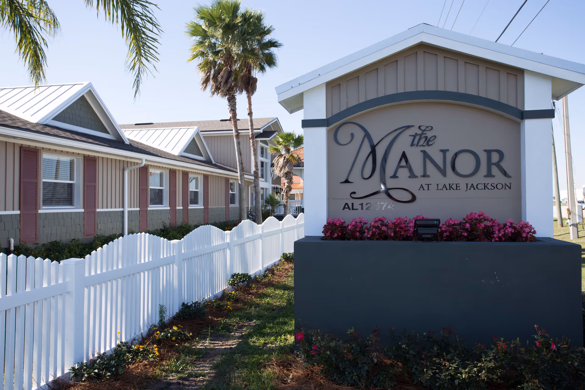 Exterior view of The Manor at Lake Jackson facility sign with flowers at its base, a white picket fence, palm trees, and a building with multiple windows in the background under a clear sky.