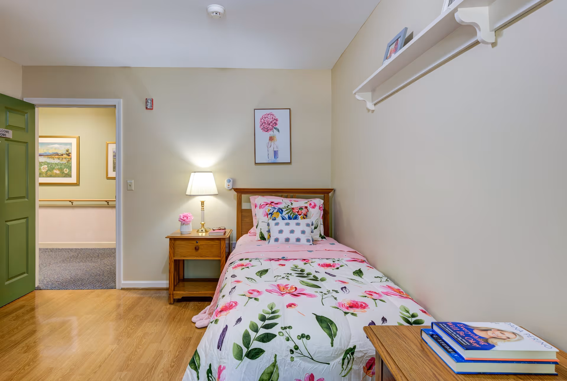 A small bedroom with a single bed covered in floral bedding. Next to the bed is a wooden nightstand with a lamp and a small flower vase. The walls are beige with a framed floral picture above the bed. A green door is open, showing a hallway with framed pictures. A wooden table with books is partially visible in the foreground.
