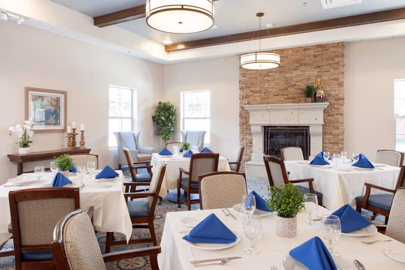 Bright dining room with tables set with white linens and blue napkins, surrounded by chairs and featuring a stone fireplace.