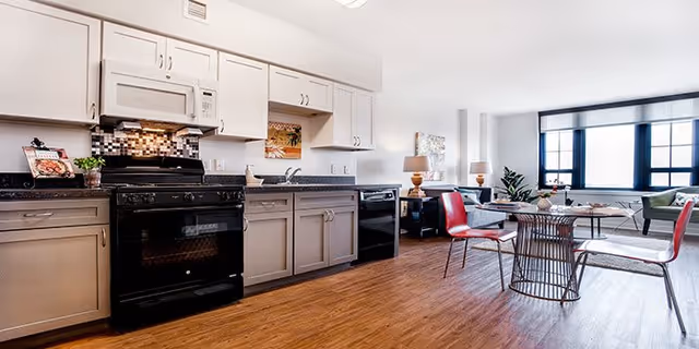 A bright and modern kitchen area with light gray cabinets, a black stove, microwave, and dishwasher. The kitchen opens into a living space with a round glass dining table surrounded by red and white chairs. The living area has large windows letting in natural light, a sofa, and decorative lamps on side tables.