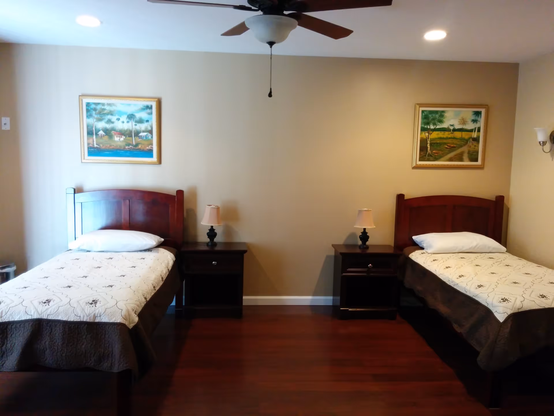 A bedroom with two single beds, each with a white pillow and patterned bedspread. Between the beds are two dark wooden nightstands, each with a small lamp. Above each bed hangs a framed landscape painting. The room has wooden flooring, beige walls, a ceiling fan with a light, and recessed ceiling lights.