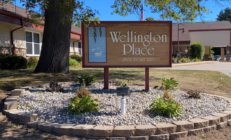 Outdoor view of the entrance area of Wellington Place at Whiting, featuring a wooden sign with the facility name and address 1902 Post Rd, surrounded by a landscaped bed with rocks and plants, with part of the building and a large tree in the background.