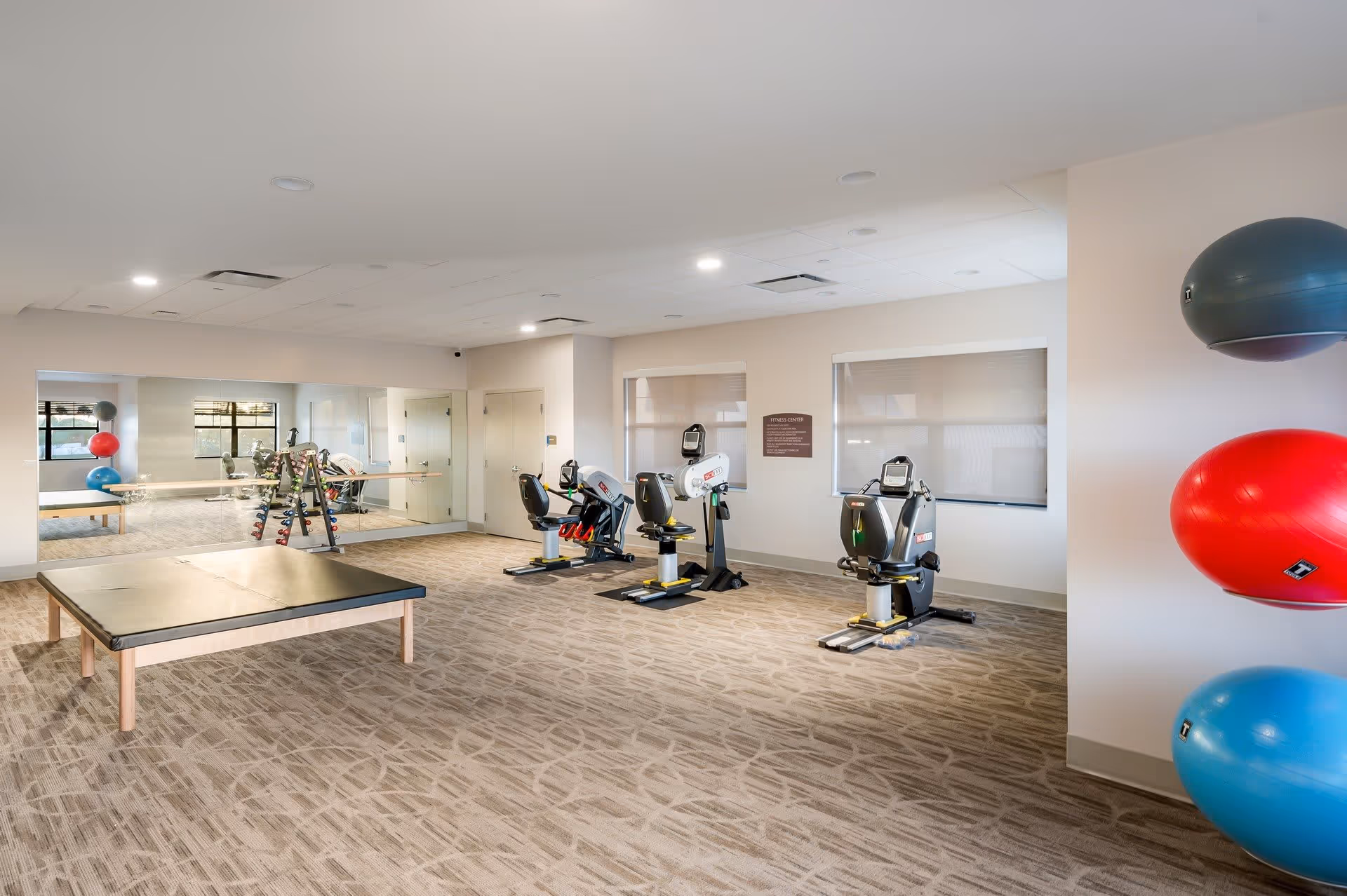 A bright fitness center room with exercise equipment including three stationary recumbent bikes, a rack of dumbbells, a padded exercise table, and three large exercise balls mounted on the wall. The room has beige walls, carpeted floor, and a large mirror on one wall reflecting the equipment.