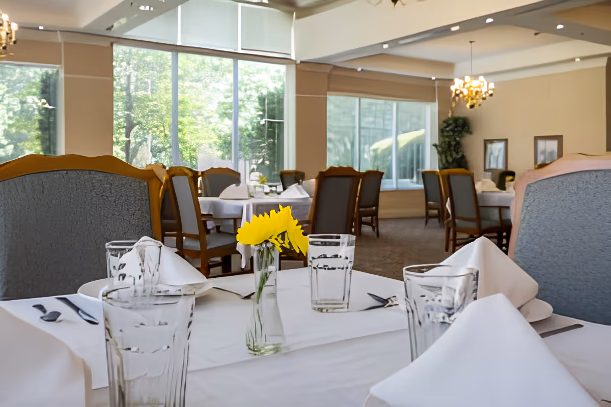 A dining room with tables covered in white tablecloths, set with glasses, napkins, and silverware. A small vase with yellow flowers is placed on the table in the foreground. The room has large windows letting in natural light and wooden chairs with cushioned seats.