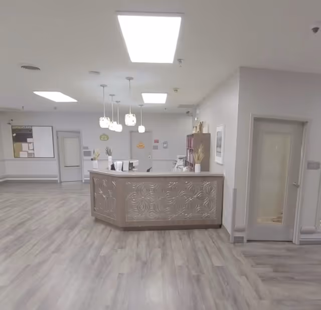 A clean and modern reception desk area inside a senior living facility with decorative paneling on the desk, pendant lights hanging from the ceiling, and light wood flooring. The walls are light-colored with a door and bulletin board visible in the background.