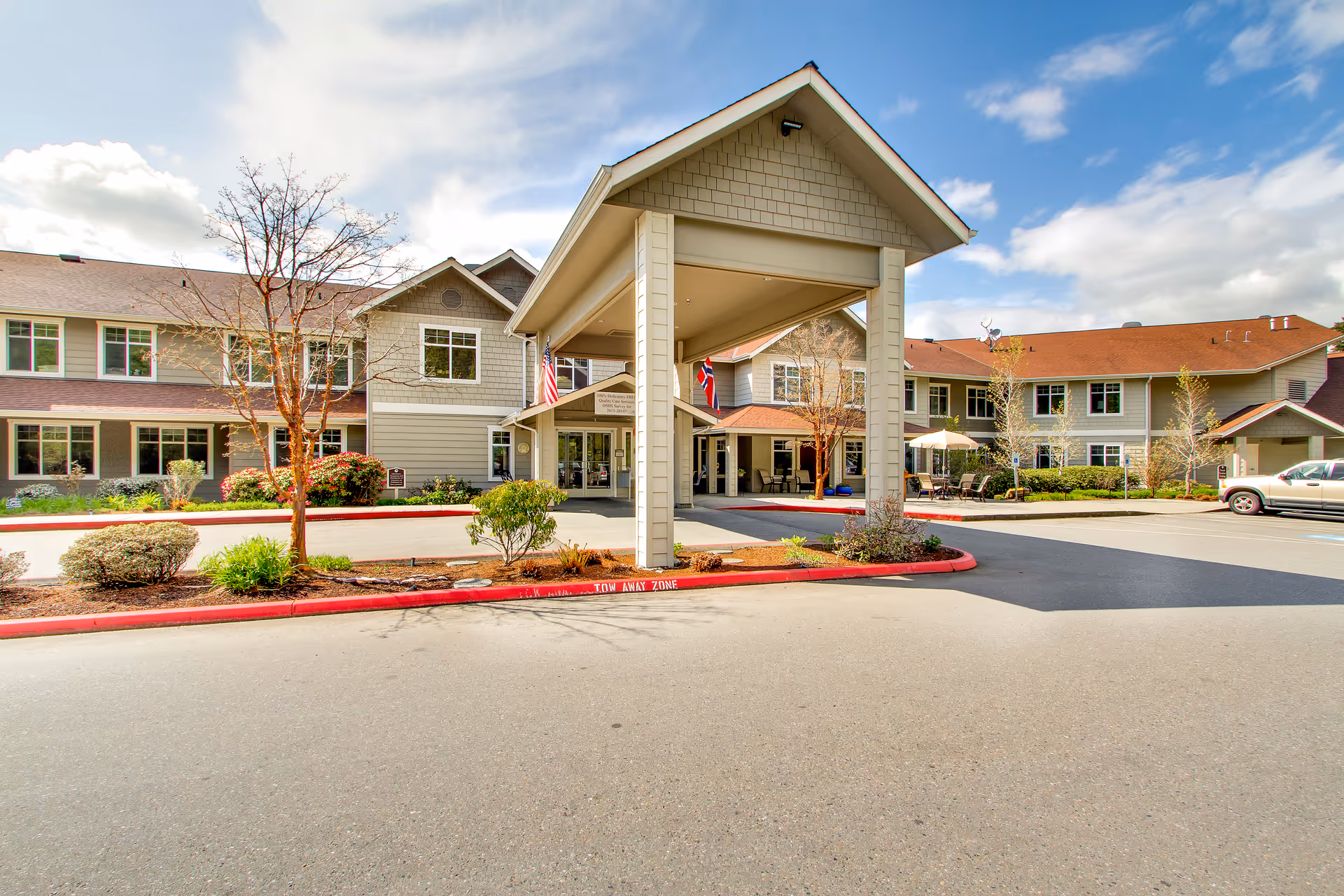 Front entrance of a two-story senior living building with a covered porte-cochere, landscaping, and parked cars.