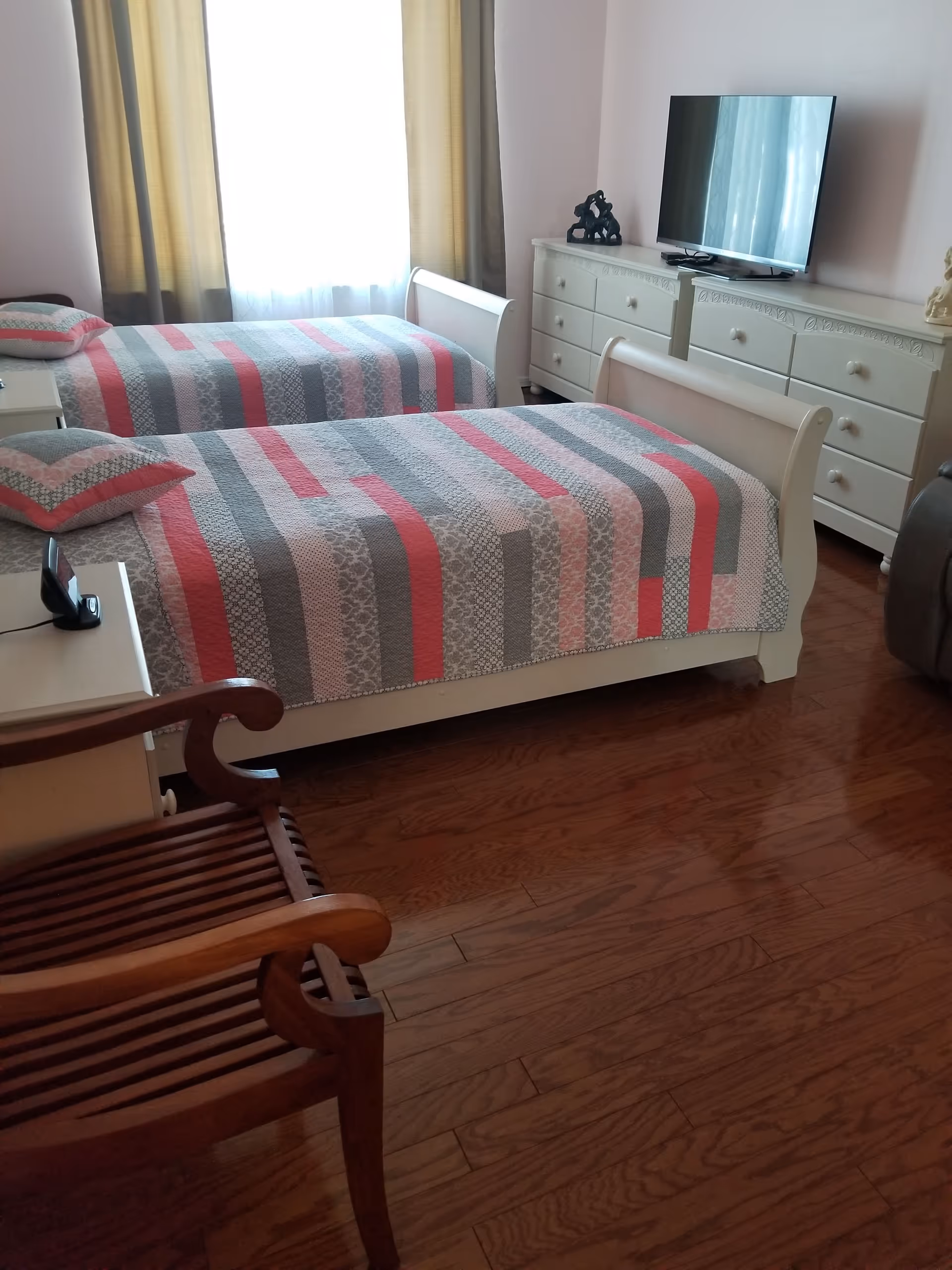 A bedroom with two twin beds covered in striped quilts in shades of pink, gray, and white. There is a wooden chair in the foreground, a white dresser with a flat-screen TV on top, and a window with beige curtains letting in natural light.