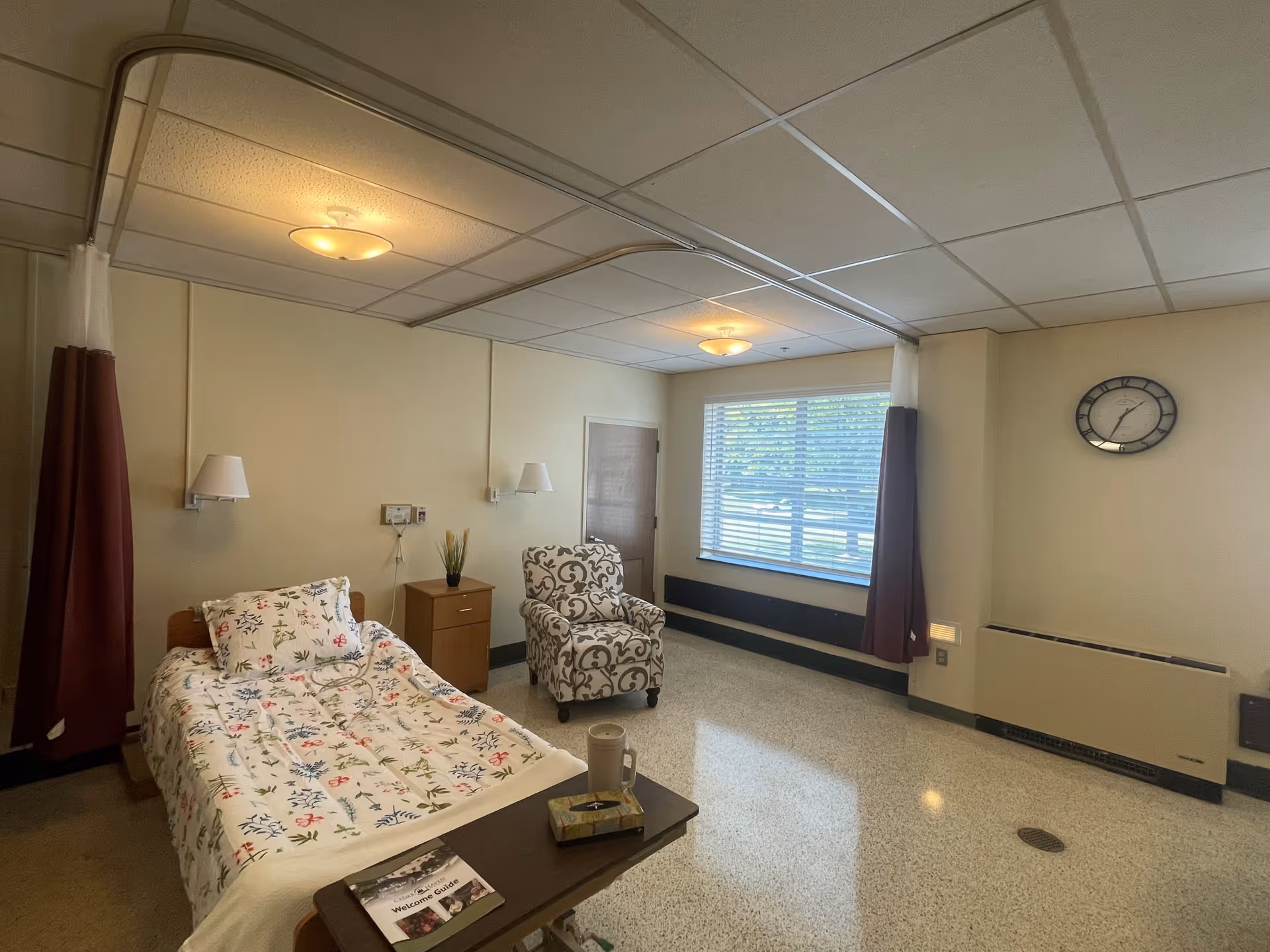 A well-lit senior living bedroom with a single bed covered in a floral-patterned blanket, a small wooden nightstand with a plant, a patterned armchair, a large window with blinds and maroon curtains, a wall clock, and a heating unit beneath the window. The room has beige walls and a tiled floor.