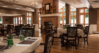 Spacious dining room with set tables and chairs, a central brick fireplace, chandeliers, and large windows overlooking greenery.