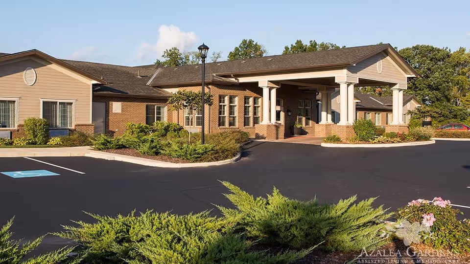 Exterior view of Azalea Gardens Assisted Living & Memory Care building with a covered entrance, brick and beige siding, surrounded by landscaped bushes and trees under a clear sky.