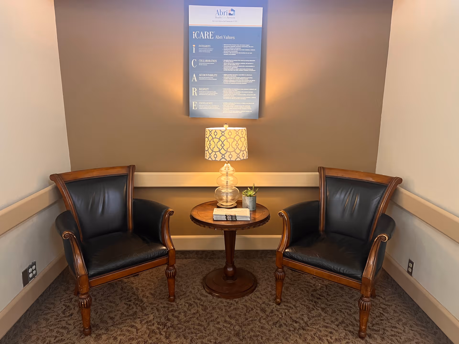 A small seating area with two wooden armchairs with black cushions facing each other and a round wooden table between them. On the table is a decorative lamp with a patterned shade, a small potted plant, and a book. Above the table on the wall is a framed poster titled 'iCARE Abri Values' with text describing values such as Integrity, Collaboration, Accountability, Respect, and Excellence.