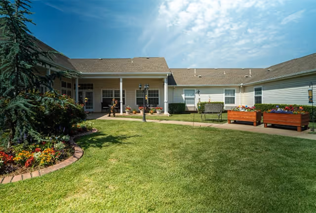 Outdoor courtyard area of a senior living facility with green grass, flower beds, benches, and a building with white siding and a covered patio under a partly cloudy sky.