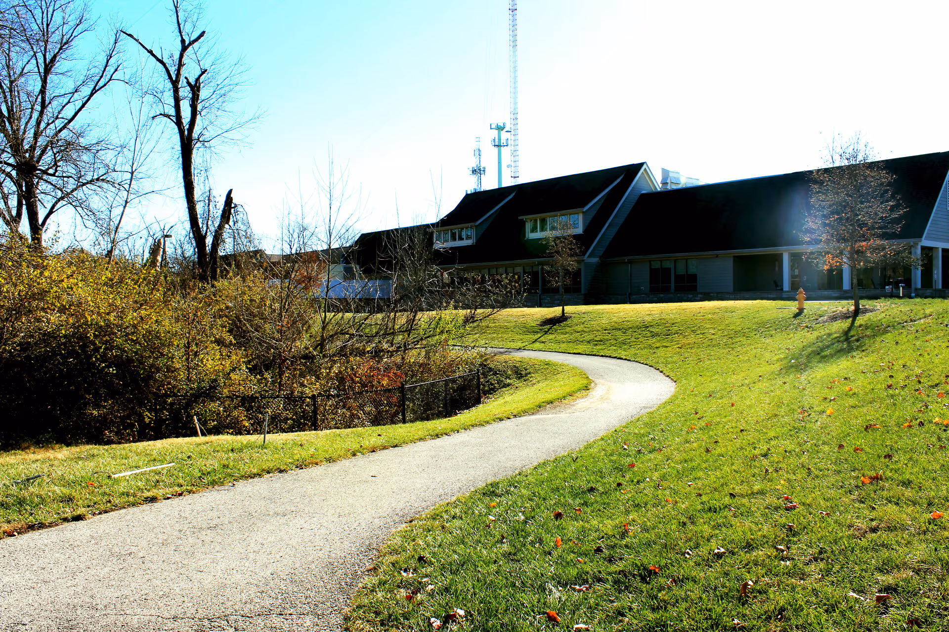 A winding paved path curves through a sunny grassy lawn toward a low building with dark roofs and bare trees.
