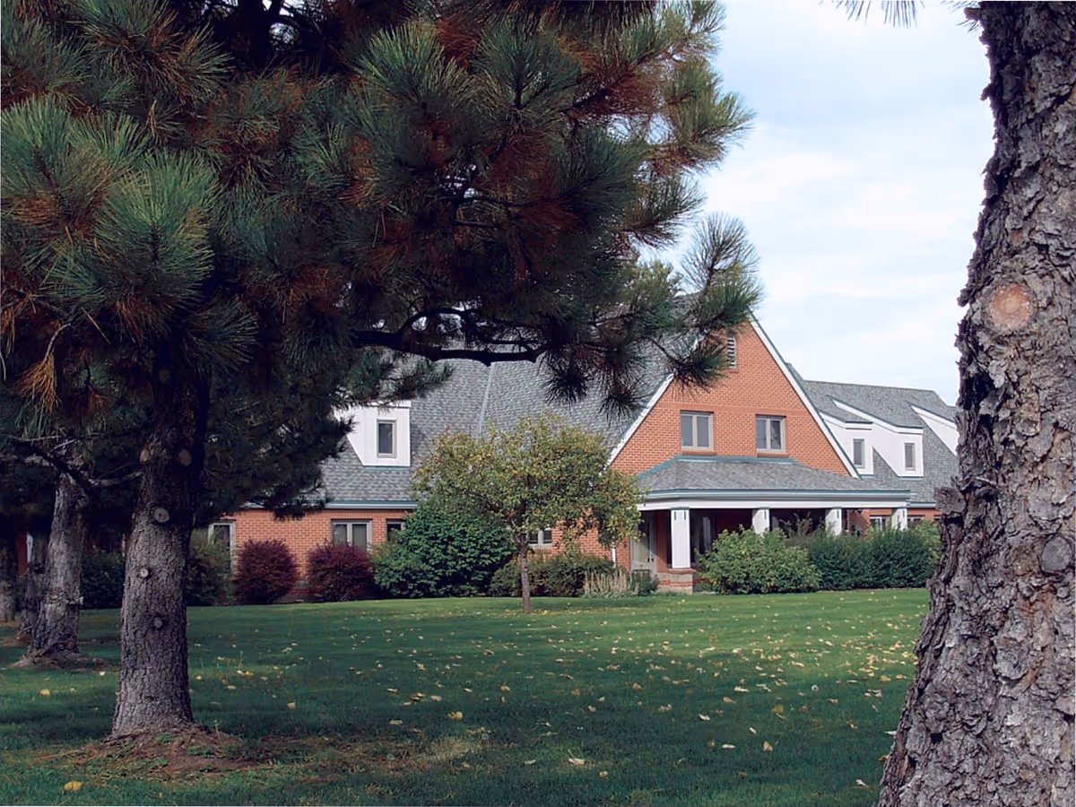 A large brick building with a steeply pitched roof and multiple windows, partially obscured by tall pine trees and other greenery in the foreground. The building is surrounded by a well-maintained lawn with scattered fallen leaves under a partly cloudy sky.