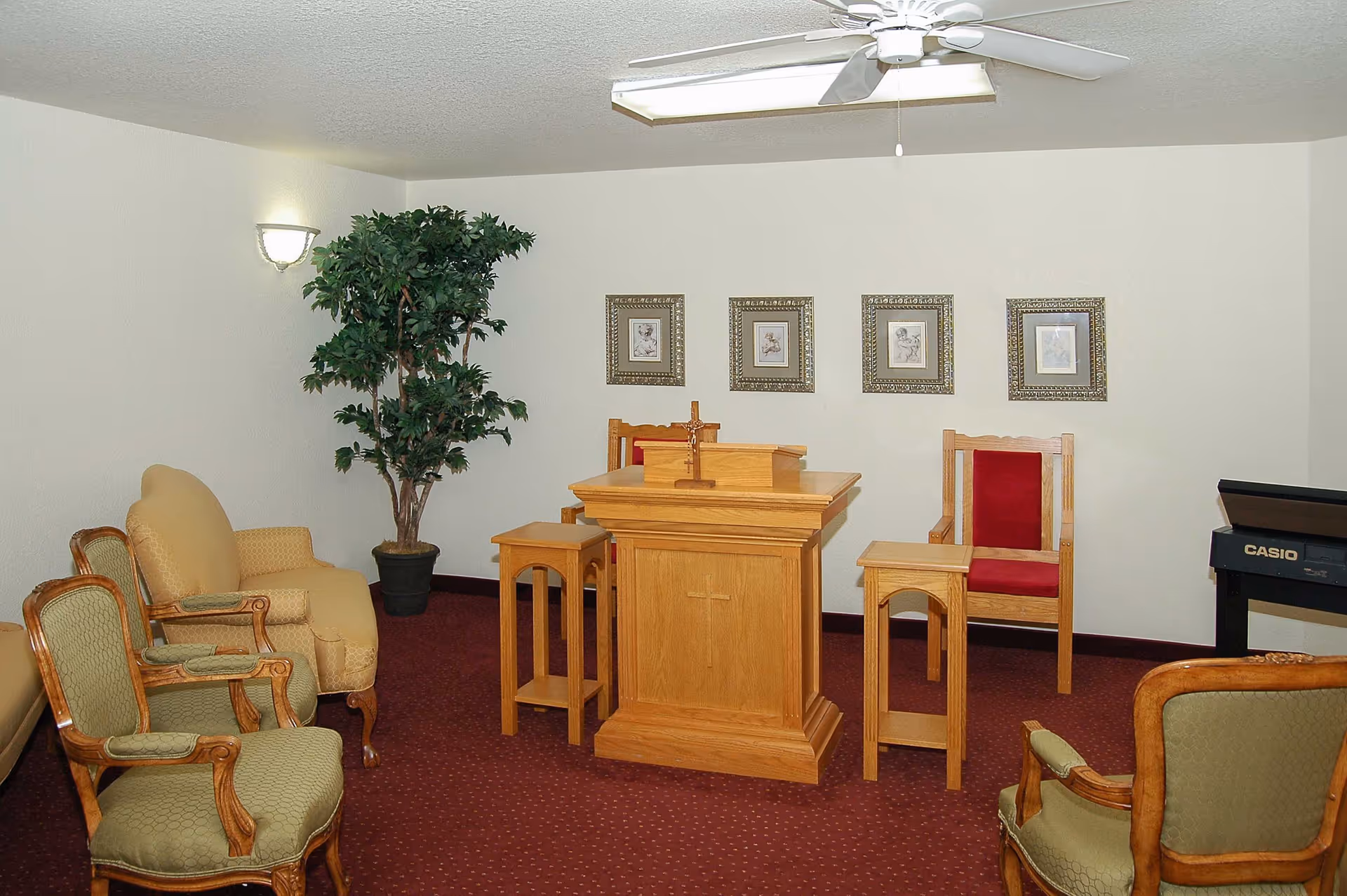 A small chapel or prayer room with a wooden pulpit featuring a cross, surrounded by several chairs including green upholstered armchairs and wooden chairs with red cushions. There is a potted plant in the corner, four framed pictures on the wall, a ceiling fan, and a Casio keyboard on the right side.