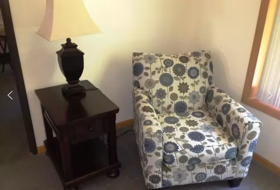 A floral patterned armchair next to a dark wooden side table with a lamp on it, placed near a window with light curtains in a room with light-colored walls and carpeted floor.