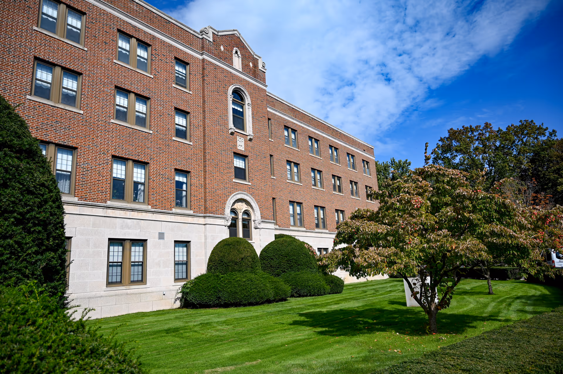 Exterior view of a multi-story brick building with numerous windows, surrounded by well-maintained green lawns, bushes, and trees under a partly cloudy blue sky.