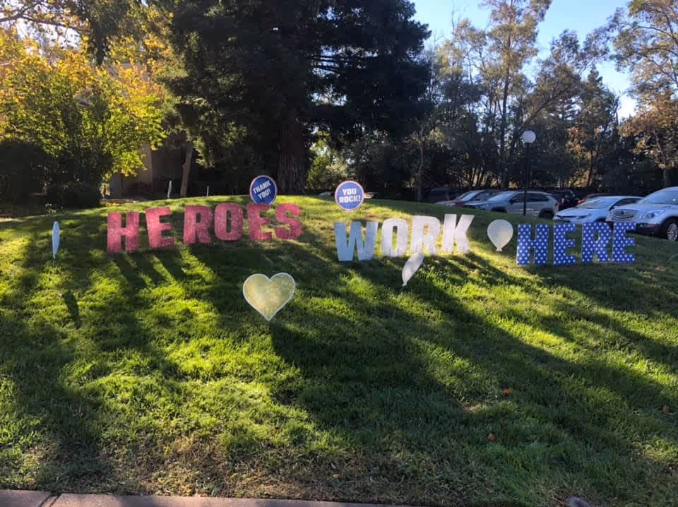 Outdoor grassy area with large colorful letters spelling out 'HEROES WORK HERE' along with heart and balloon shapes. Two circular signs say 'THANK YOU!' and 'YOU ROCK!'. Trees and parked cars are visible in the background under a clear sky.