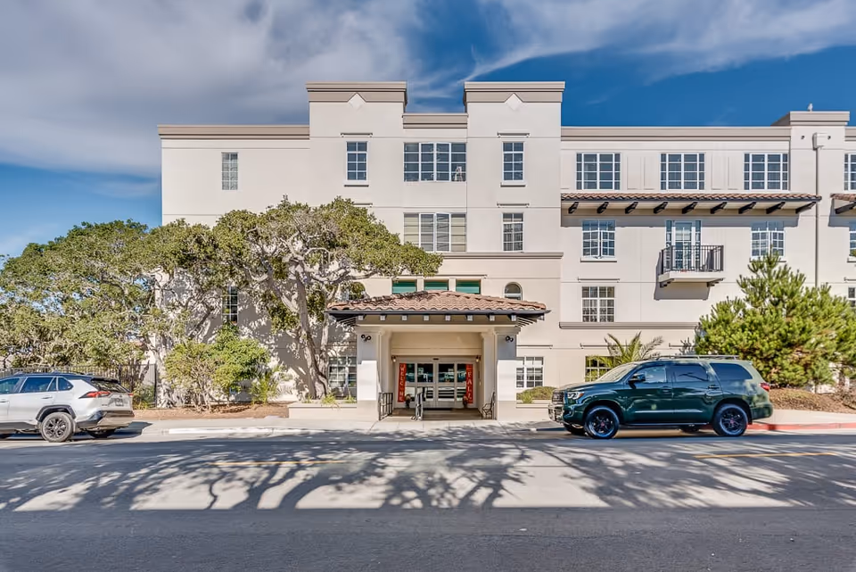 Front exterior view of a multi-story senior living facility building with a covered entrance, several windows, and two parked cars on the street in front. There are trees and landscaping around the building under a partly cloudy sky.
