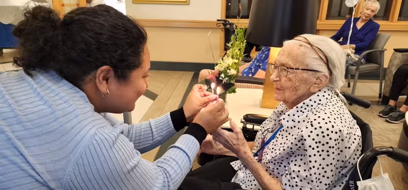 A young woman lighting a candle held by an elderly woman sitting in a wheelchair inside a common area of a senior living facility. Another elderly woman is seated in the background, smiling and observing the scene.