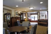 A cozy living room area with a round wooden table surrounded by upholstered chairs. The room features a fireplace with a mantel, a grandfather clock, framed artwork on the walls, and a large window letting in natural light. There are additional seating options including a bench and armchairs, with carpeted flooring and ceiling lights.