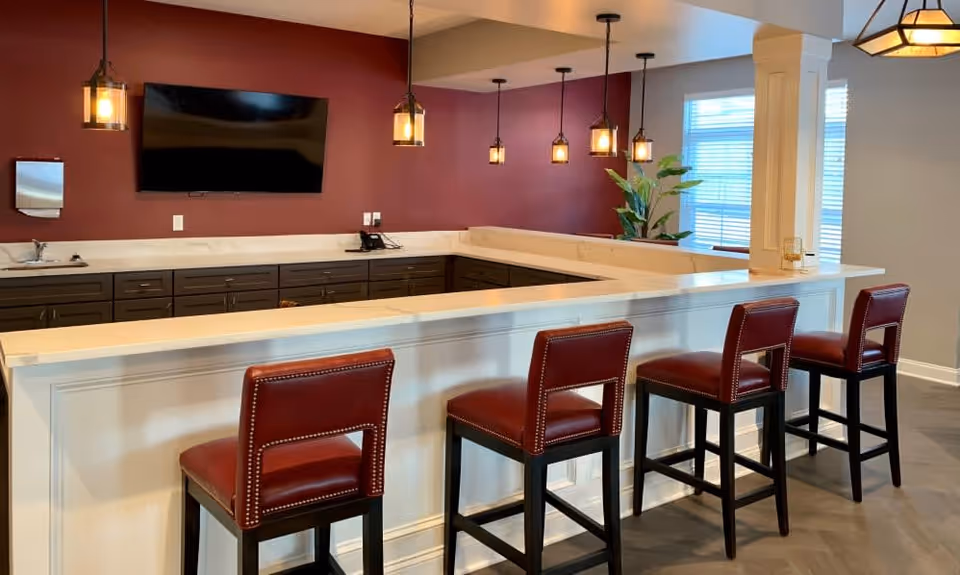 Interior view of a modern bar area with a white countertop and four red leather bar stools. The back wall is painted dark red and features a mounted flat-screen TV, a small sink, and several dark cabinets. Pendant lights hang from the ceiling above the bar, and a large window with blinds allows natural light into the space. There is also a green plant near the window.