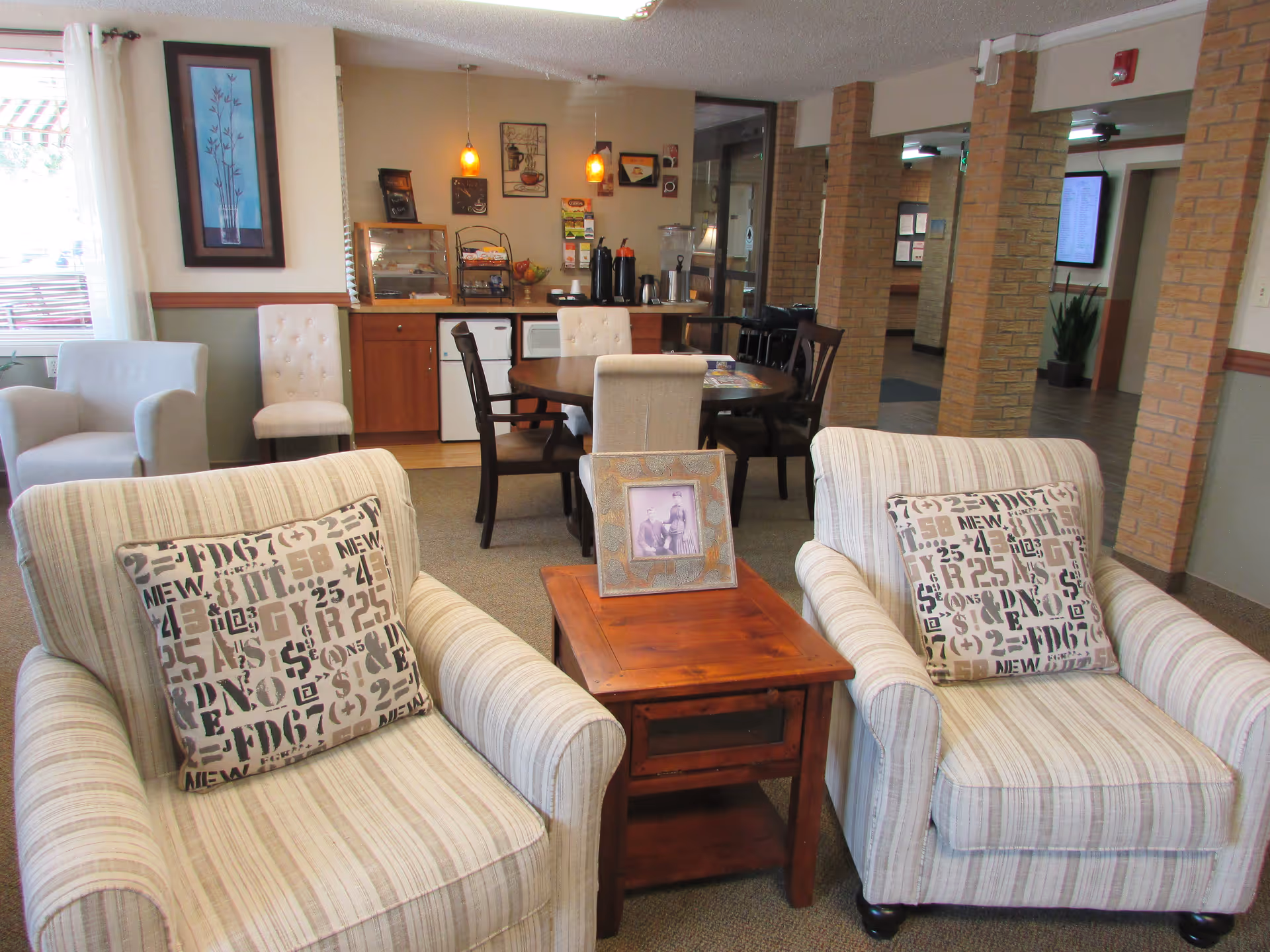Two striped armchairs with patterned pillows flank a wooden side table in a bright senior living common area with a dining table and coffee station in the background.