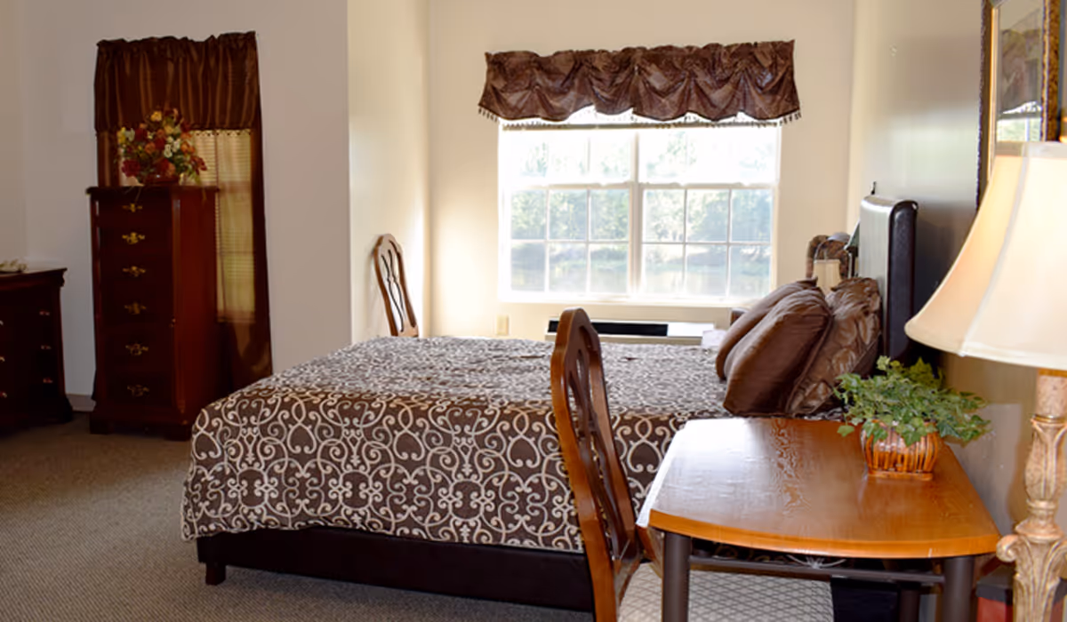 Sunlit furnished bedroom with a patterned bed, wooden dressers, a desk with lamp and chairs by a large window.