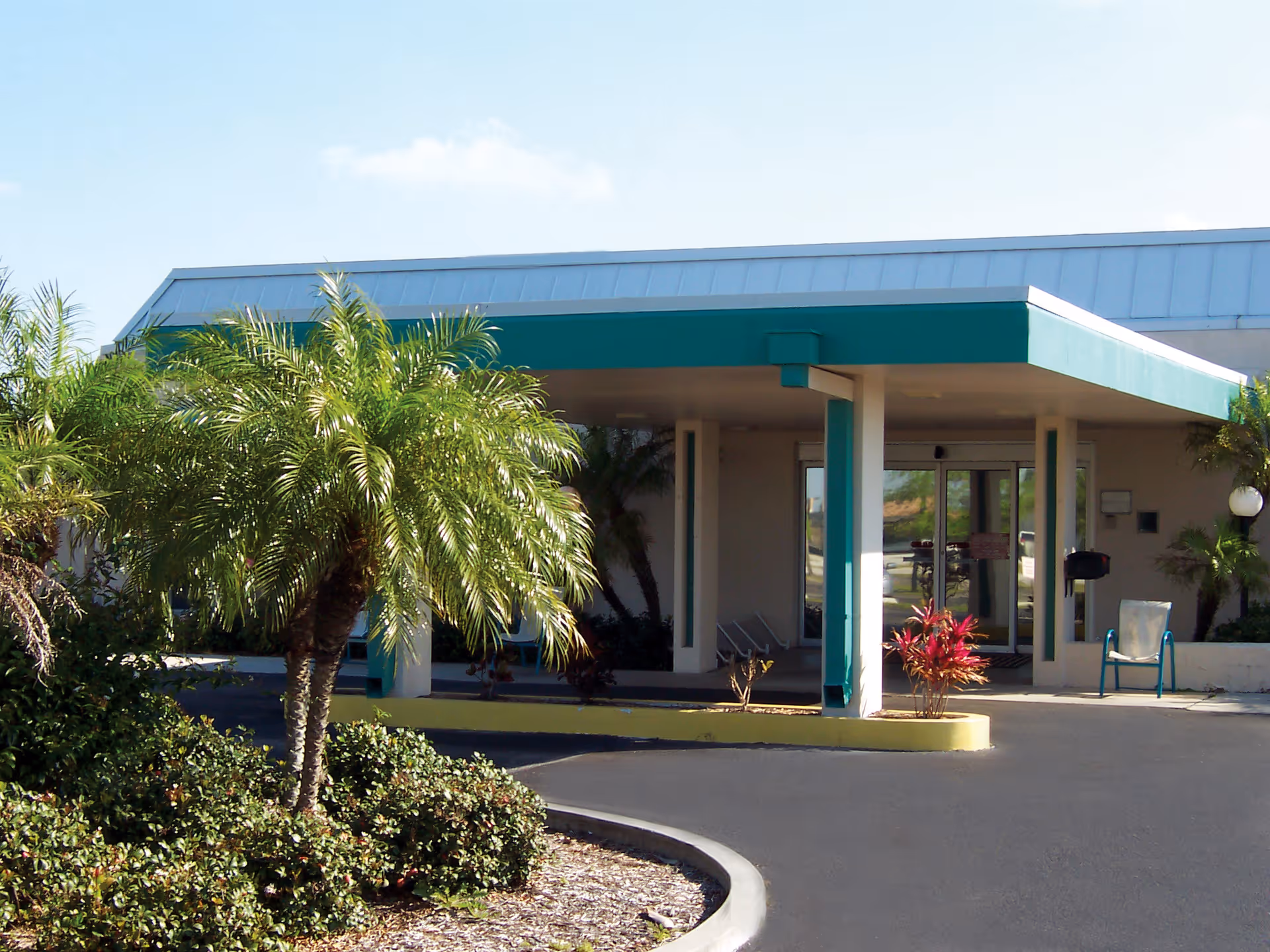 Entrance of Village Place Health and Rehabilitation Center with a covered drop-off area, surrounded by palm trees and shrubs under a clear sky.
