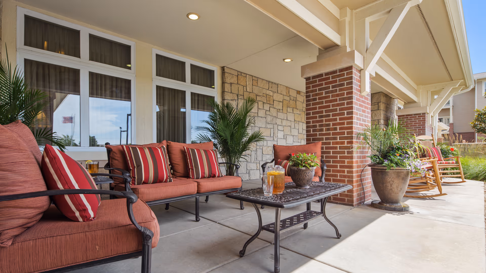 Covered porch seating area with red-cushioned patio sofas, a metal coffee table with drinks and potted plants, and rocking chairs.