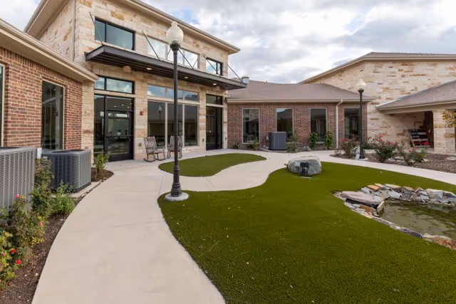 Outdoor courtyard area of Sedona Trace Health & Wellness featuring a paved walkway, green artificial turf, a small pond with rocks, lamp posts, and a building with large windows and stone and brick exterior walls.